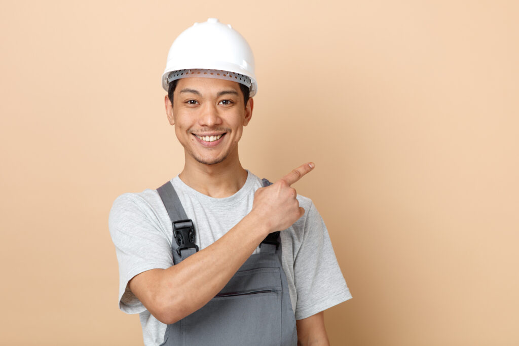 smiling young construction worker wearing safety helmet and uniform looking at camera pointing up at corner isolated on creamy background with copy space